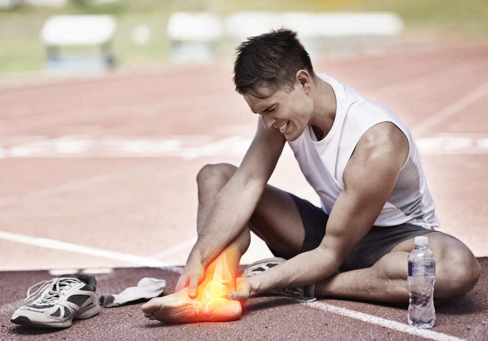 A person sitting on a running track, holding their glowing, injured ankle, with a pair of sneakers and a water bottle beside them.