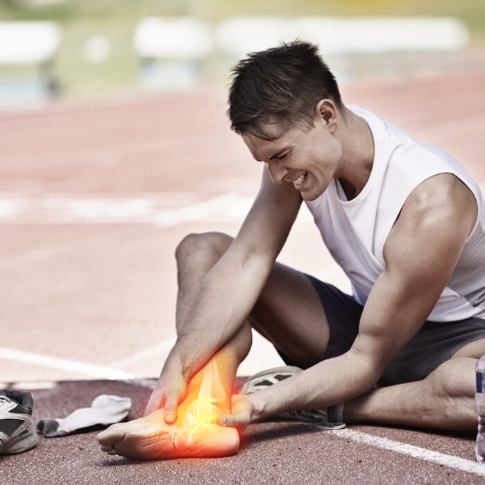 A person sitting on a running track, holding their glowing, injured ankle, with a pair of sneakers and a water bottle beside them.
